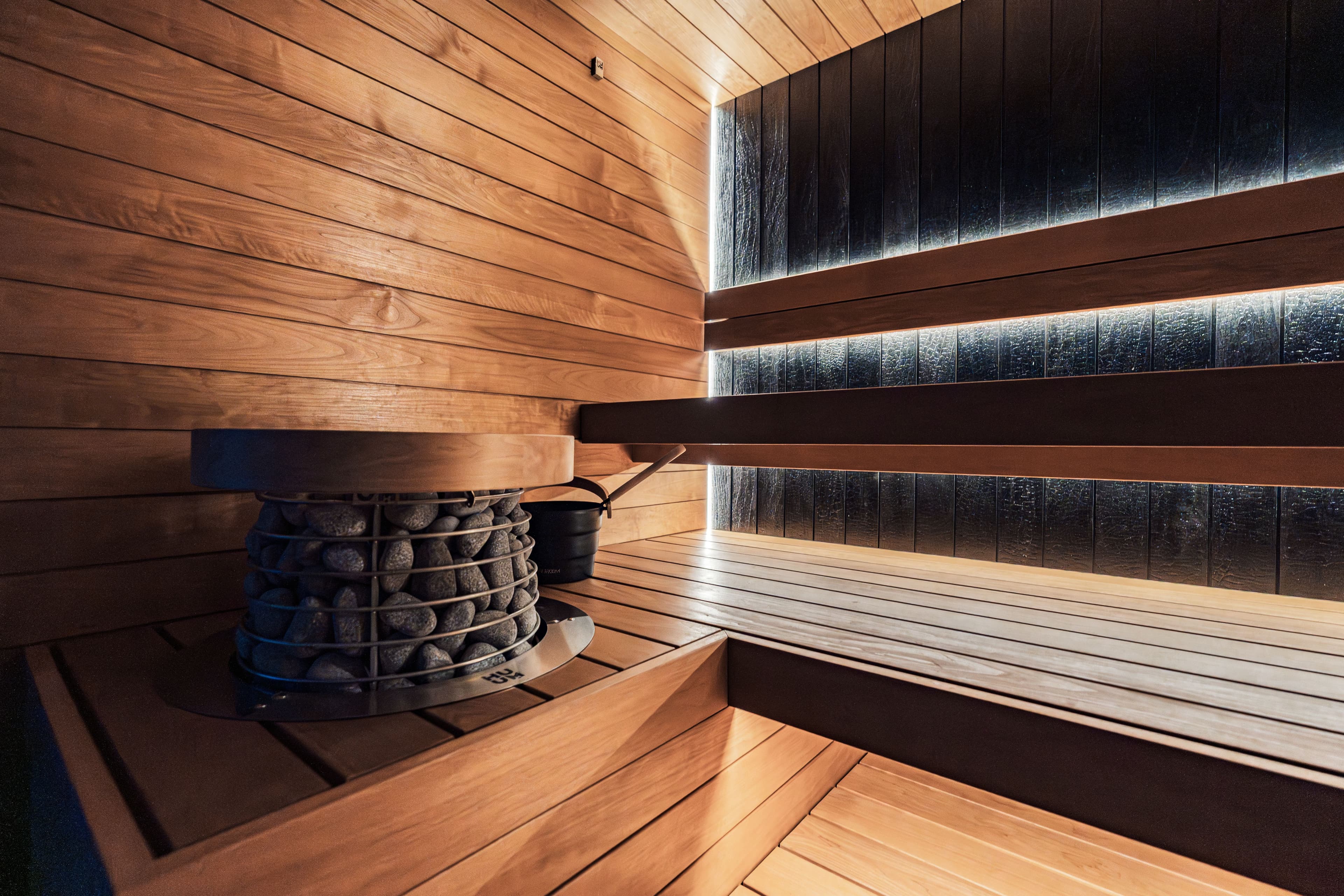 The wood-paneled interior of a sauna, with a stone heater and backlit slatted wall